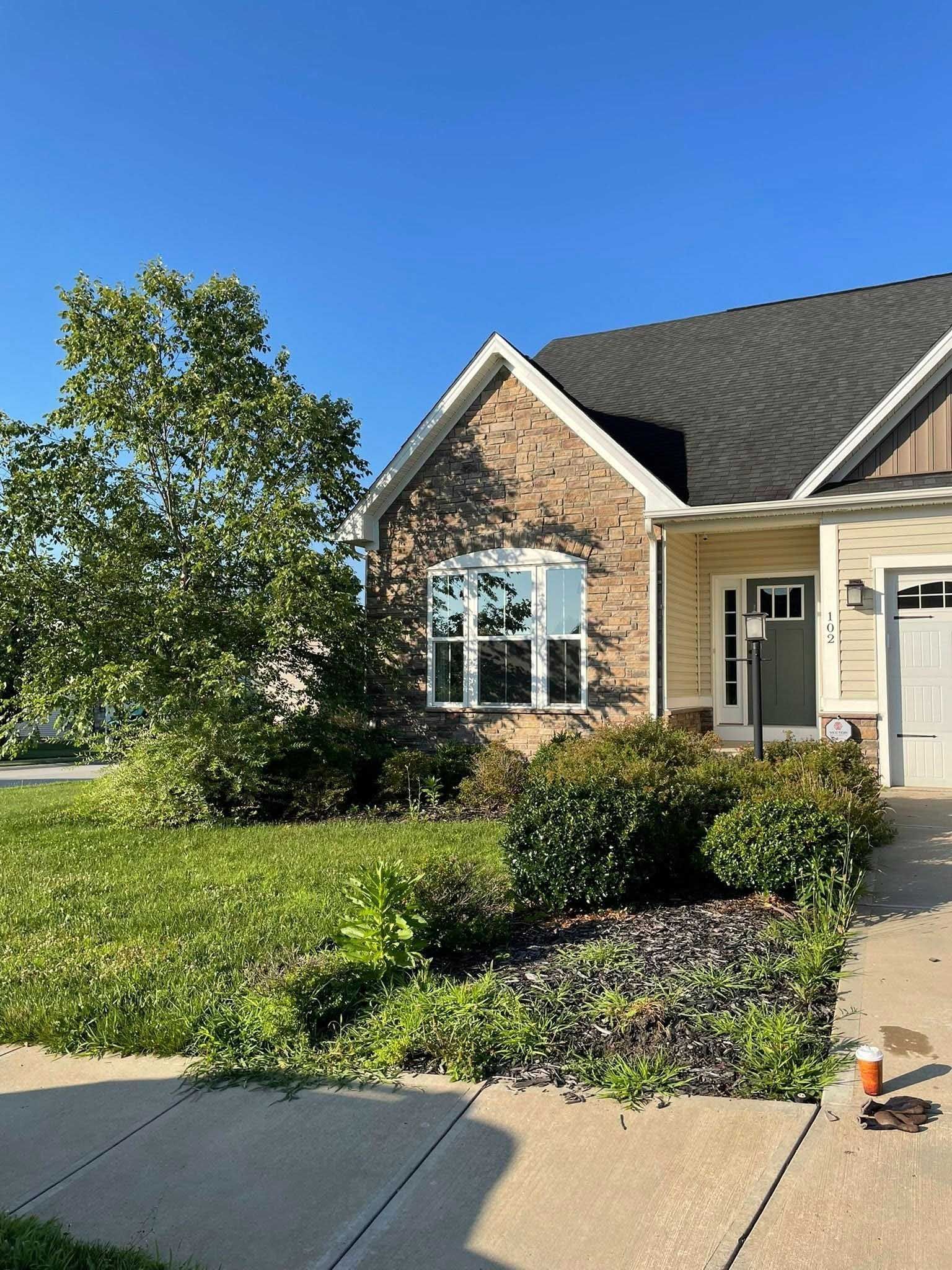 A brick and beige house with a small front yard. Bushes and a tree sit in front of the house, with a concrete sidewalk in the foreground.