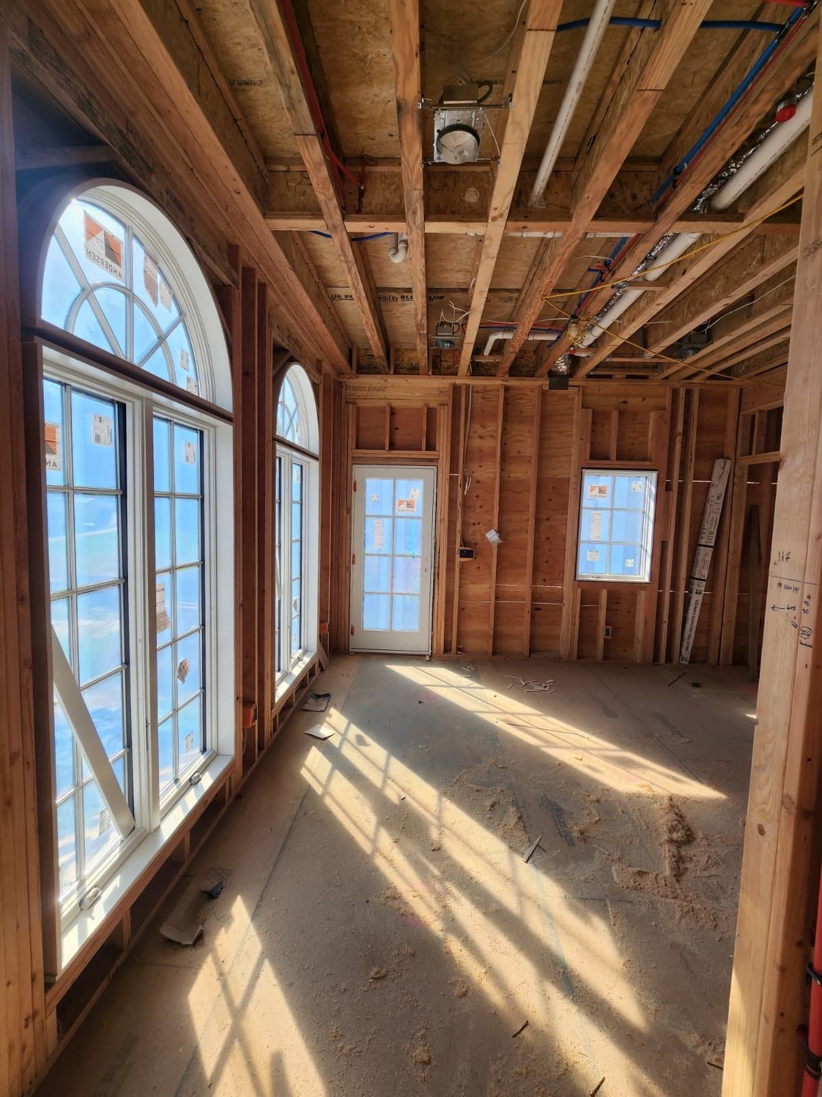 A room under construction with wood framing, exposed ceiling joists, and multiple large windows letting in sunlight.