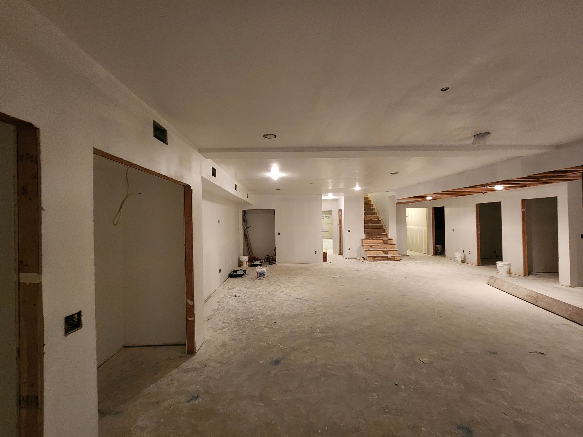 A wide-angle view of a large, unfinished basement space with white walls, unfinished floors, and a wooden staircase.