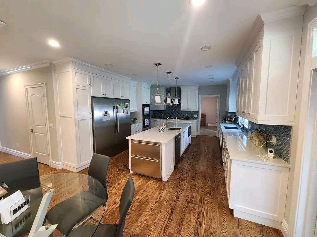 A modern white kitchen with a large central island, stainless steel appliances, and dark hardwood floors.
