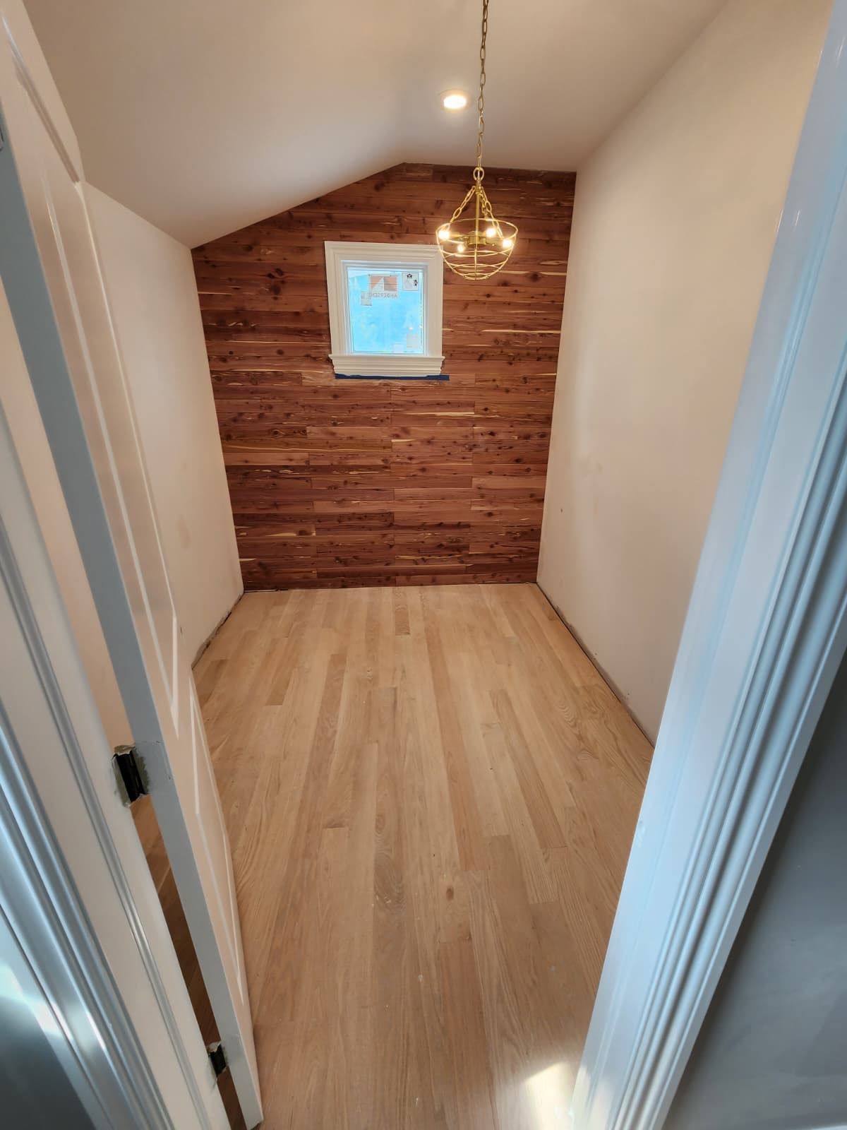 A small, empty room with light wood flooring, white walls, and a rustic wood-plank accent wall with a centered window.