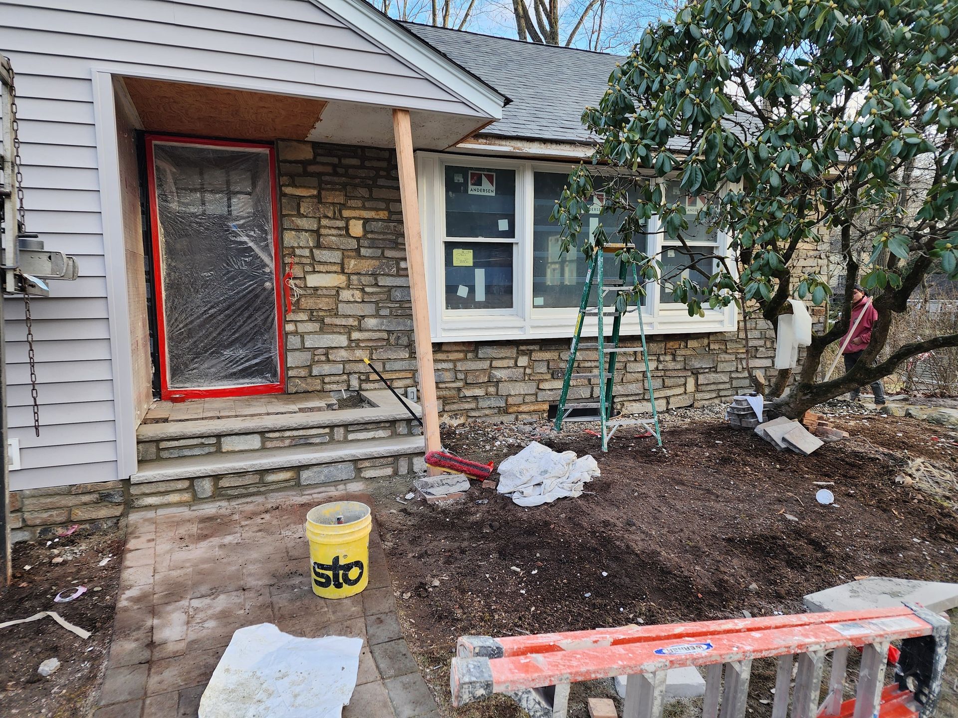 A house exterior under construction with a stone facade, a plastic-covered front door, a ladder, and a yellow bucket.