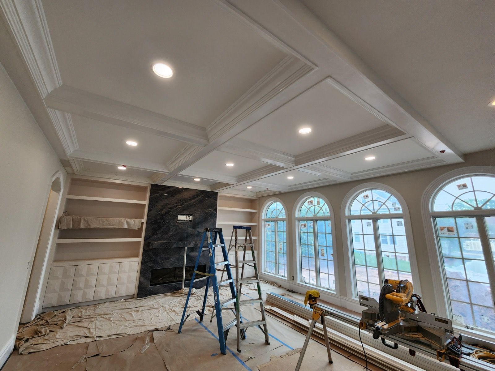 A room under construction featuring a coffered ceiling with recessed lights, a dark marble fireplace, and tall arched windows.