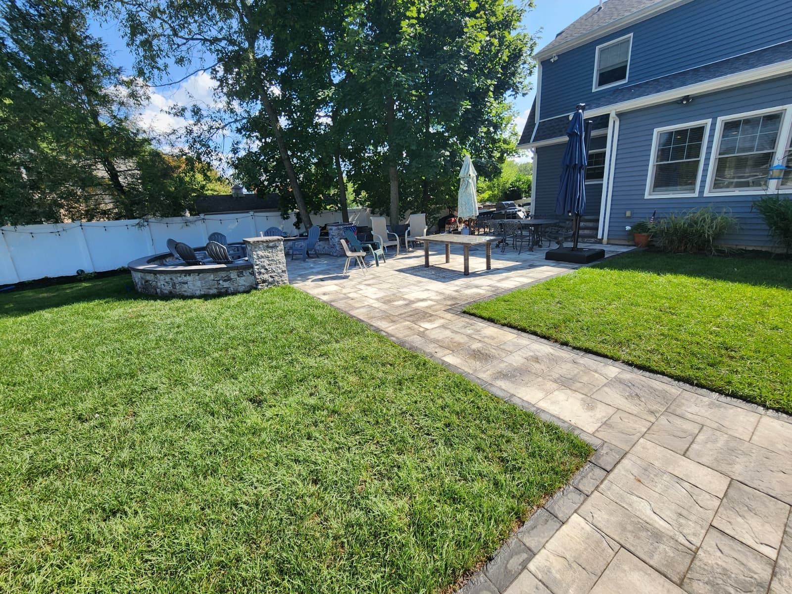 A backyard with a stone patio, fire pit area, and dining furniture, set next to a blue house and lush green lawn.