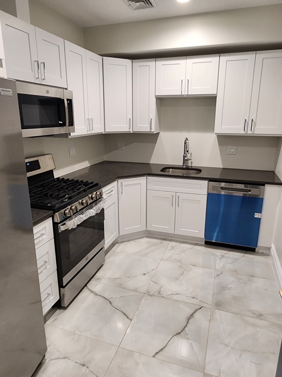Modern kitchen with white cabinets, stainless steel appliances, dark countertops, and light marble-patterned tile floors.
