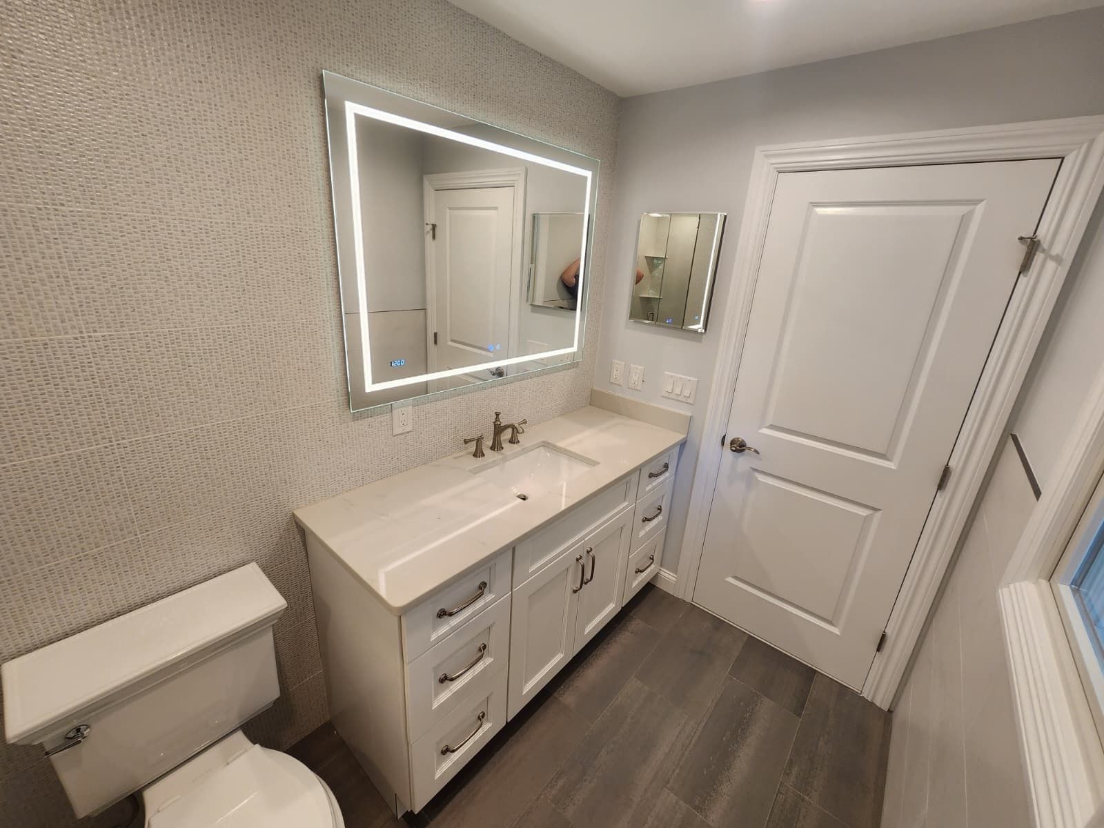 Modern bathroom vanity with a lighted rectangular mirror, white cabinets, grey wood flooring, and an adjacent door.