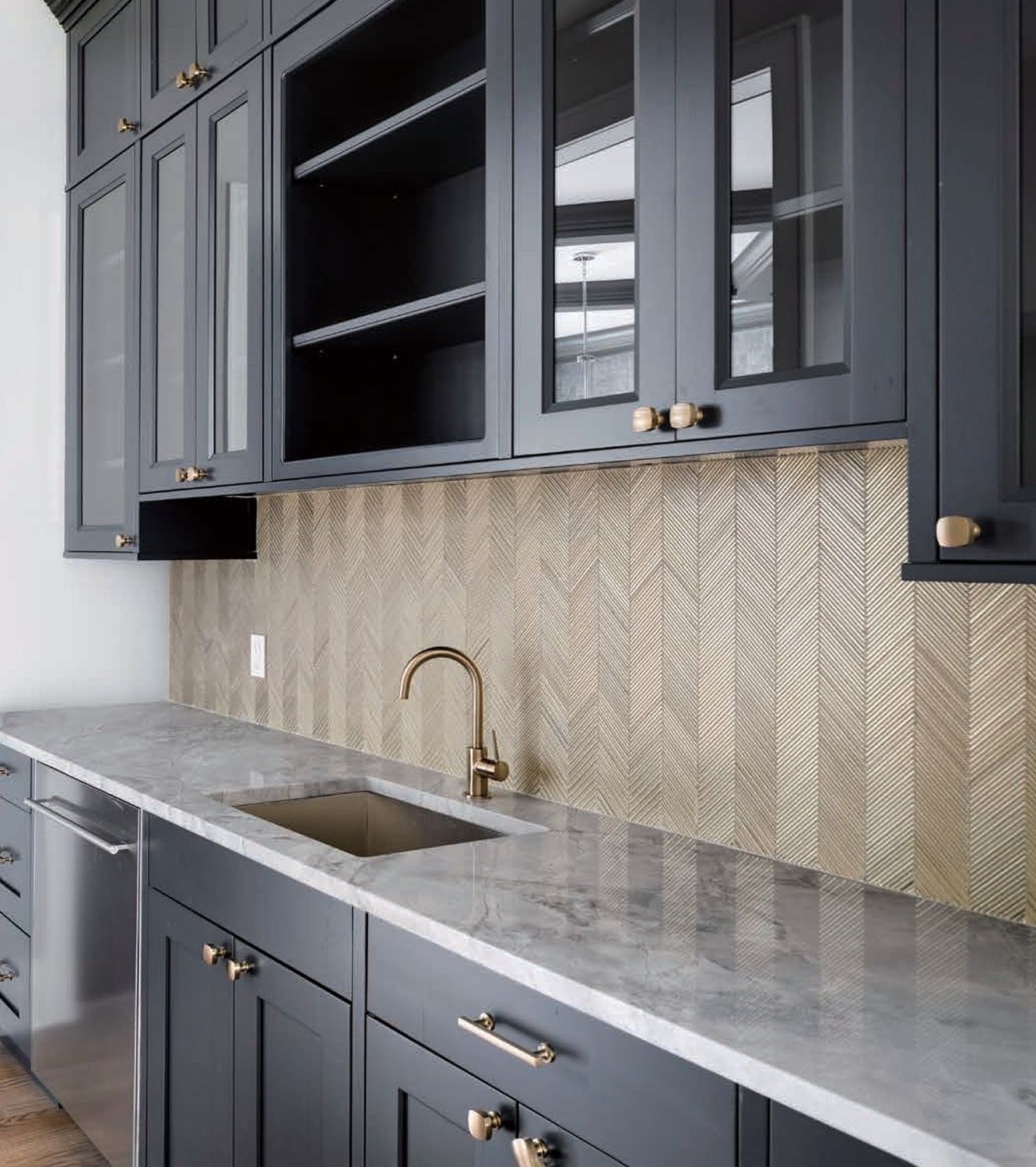A modern kitchen with dark gray cabinets, a marble countertop, gold faucet, and a metallic herringbone tile backsplash.