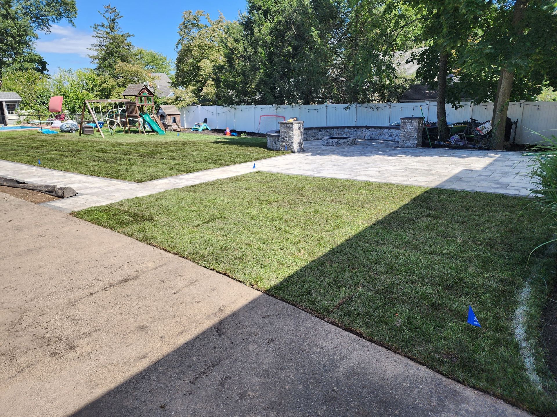 A backyard scene featuring a stone patio, newly installed sod lawns, and a wooden swing set against a white fence.