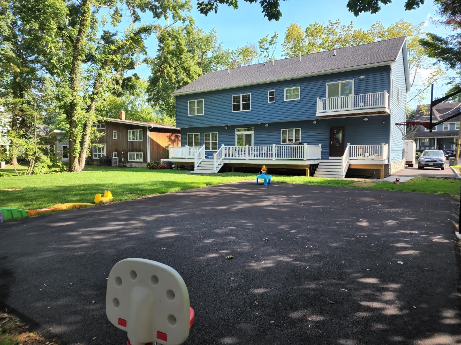 A blue house with a white deck and balcony stands behind a large paved driveway with a basketball hoop in the foreground.