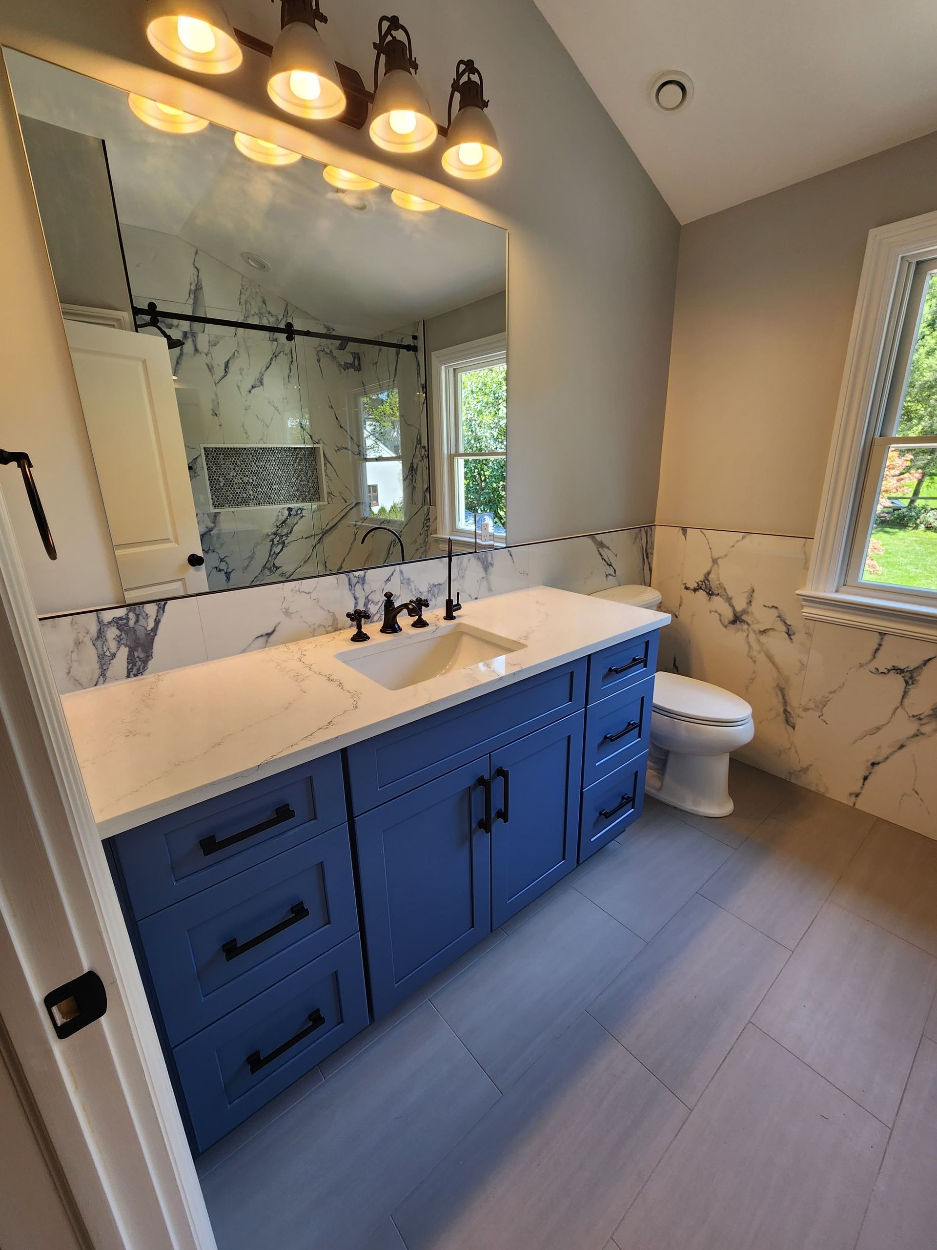 A modern bathroom with blue cabinetry, white marble countertops and wall tiles, a large mirror, and a toilet.