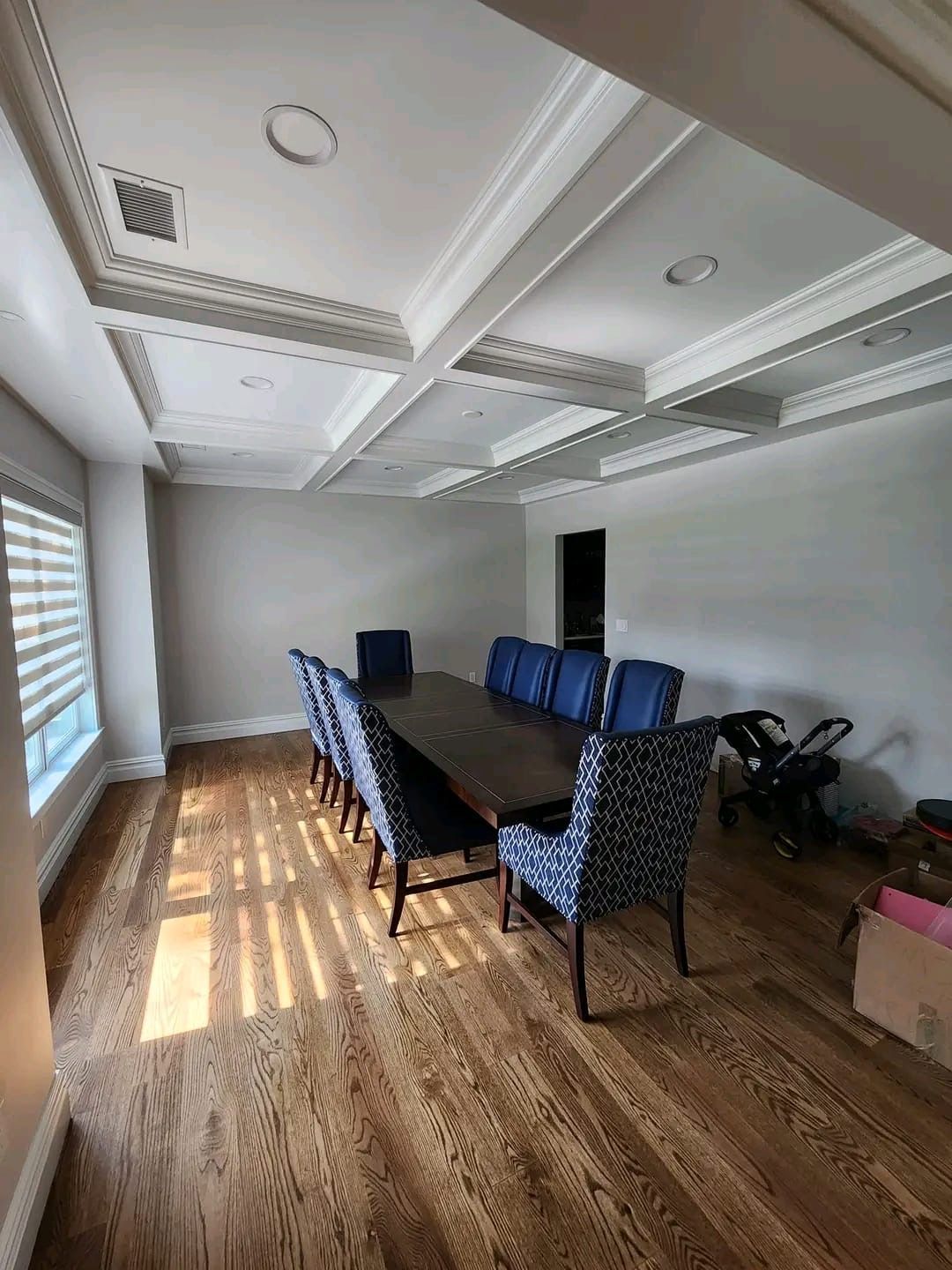 A long wooden dining table with patterned blue chairs in a bright, modern room with coffered ceilings and wood floors.