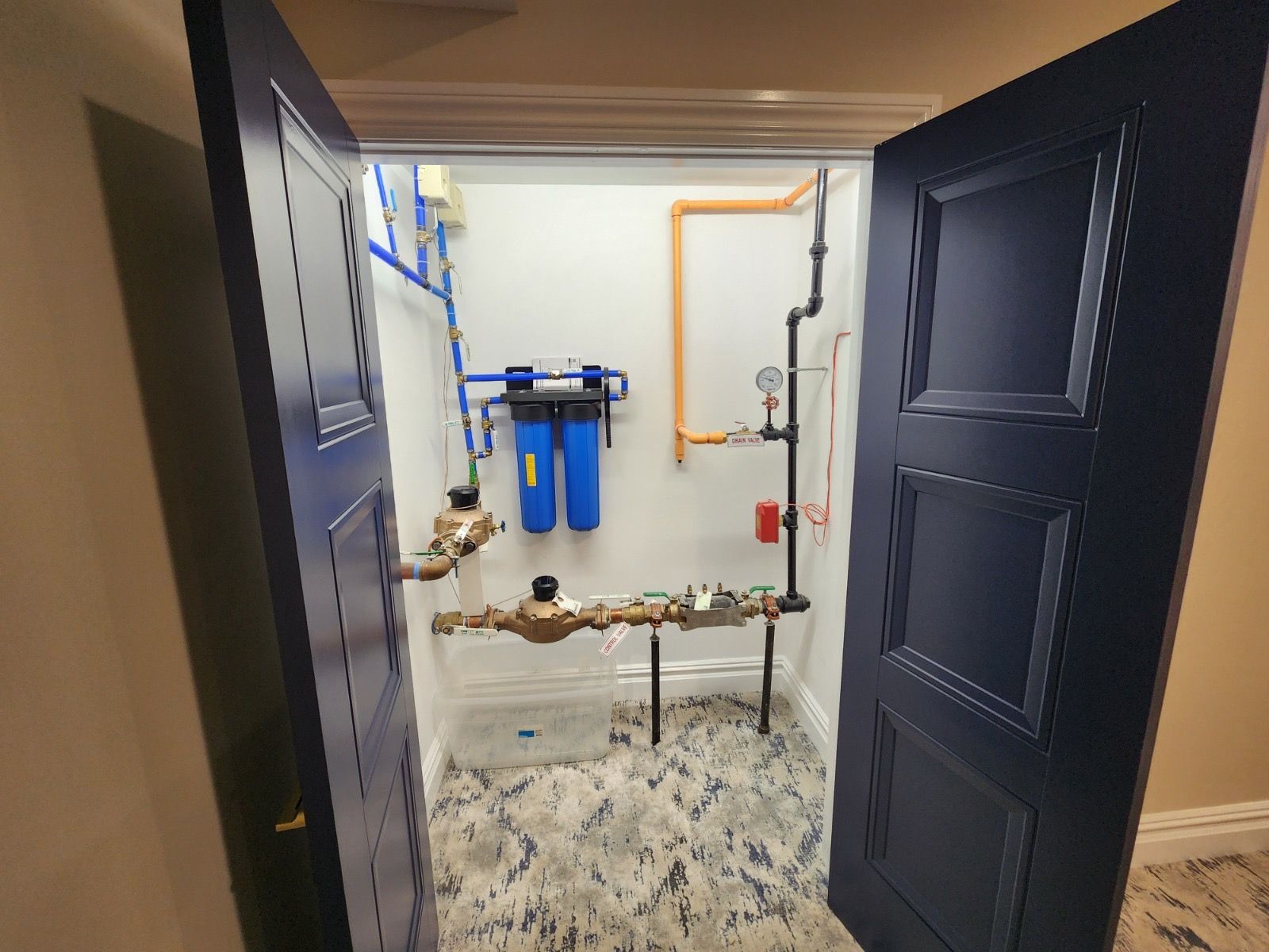 A utility closet with blue water filters, copper plumbing pipes, and a water meter against a white wall.
