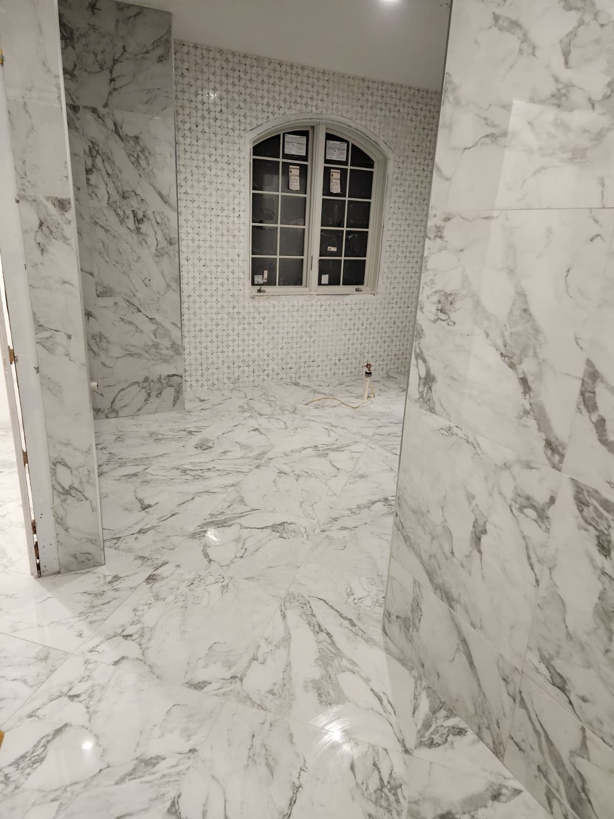 A room interior featuring white marble-patterned tile floors and walls, with a centered arched window.