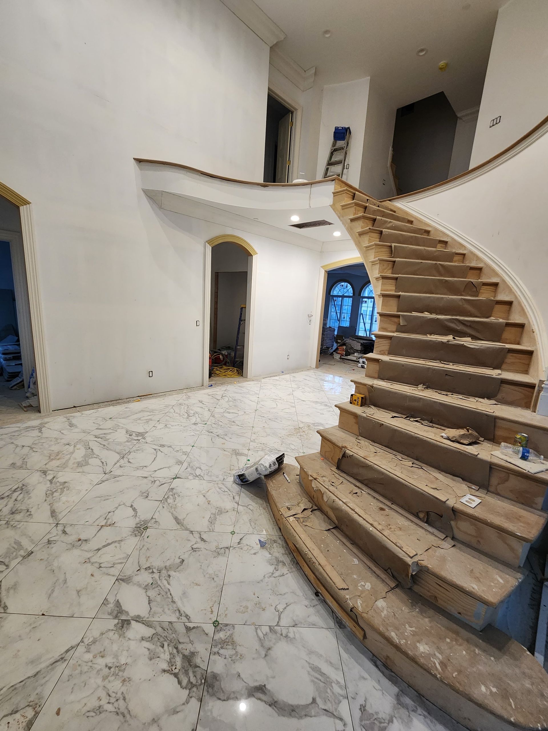 Interior of a home under construction, featuring a large marble floor, arched doorways, and an unfinished curved staircase.