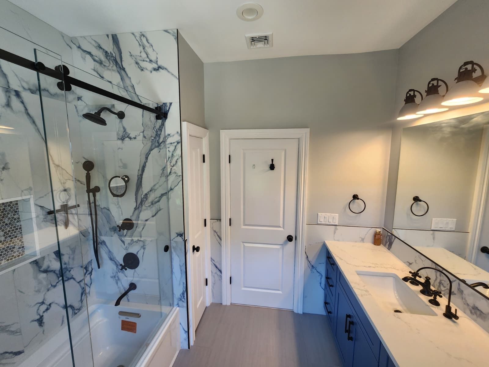 A modern bathroom with blue vanity cabinets, white marble shower walls, glass sliding doors, and a white interior door.