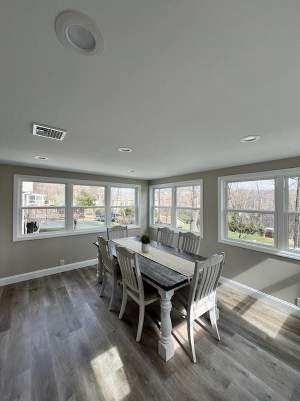 A bright, modern dining room with a long wooden table, light-colored chairs, and large windows overlooking trees.