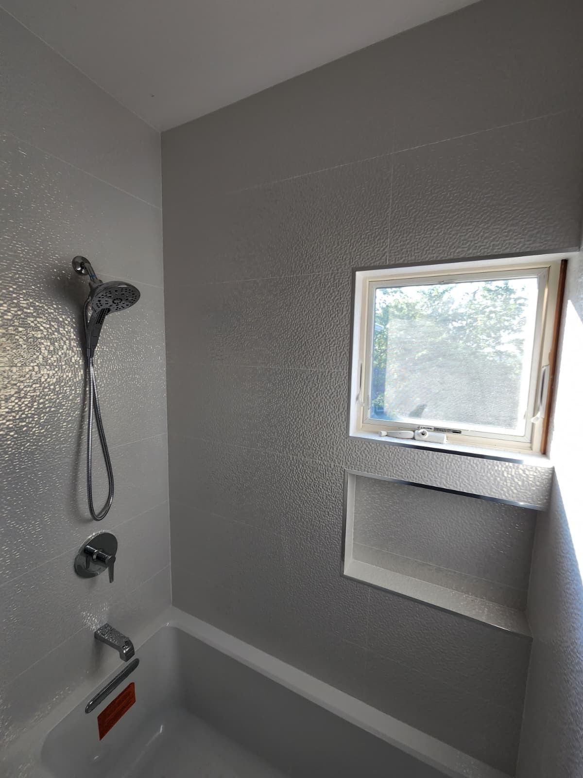 A bathroom bathtub with a shower head, faucet, and a small recessed wall niche near a window with a textured grey finish.