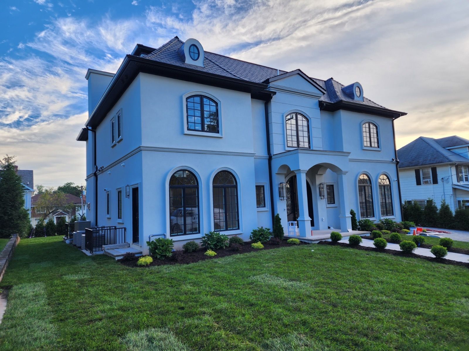 Large, blue, two-story house with arched windows, a dark shingled roof, and a grassy front lawn under a cloudy sky.
