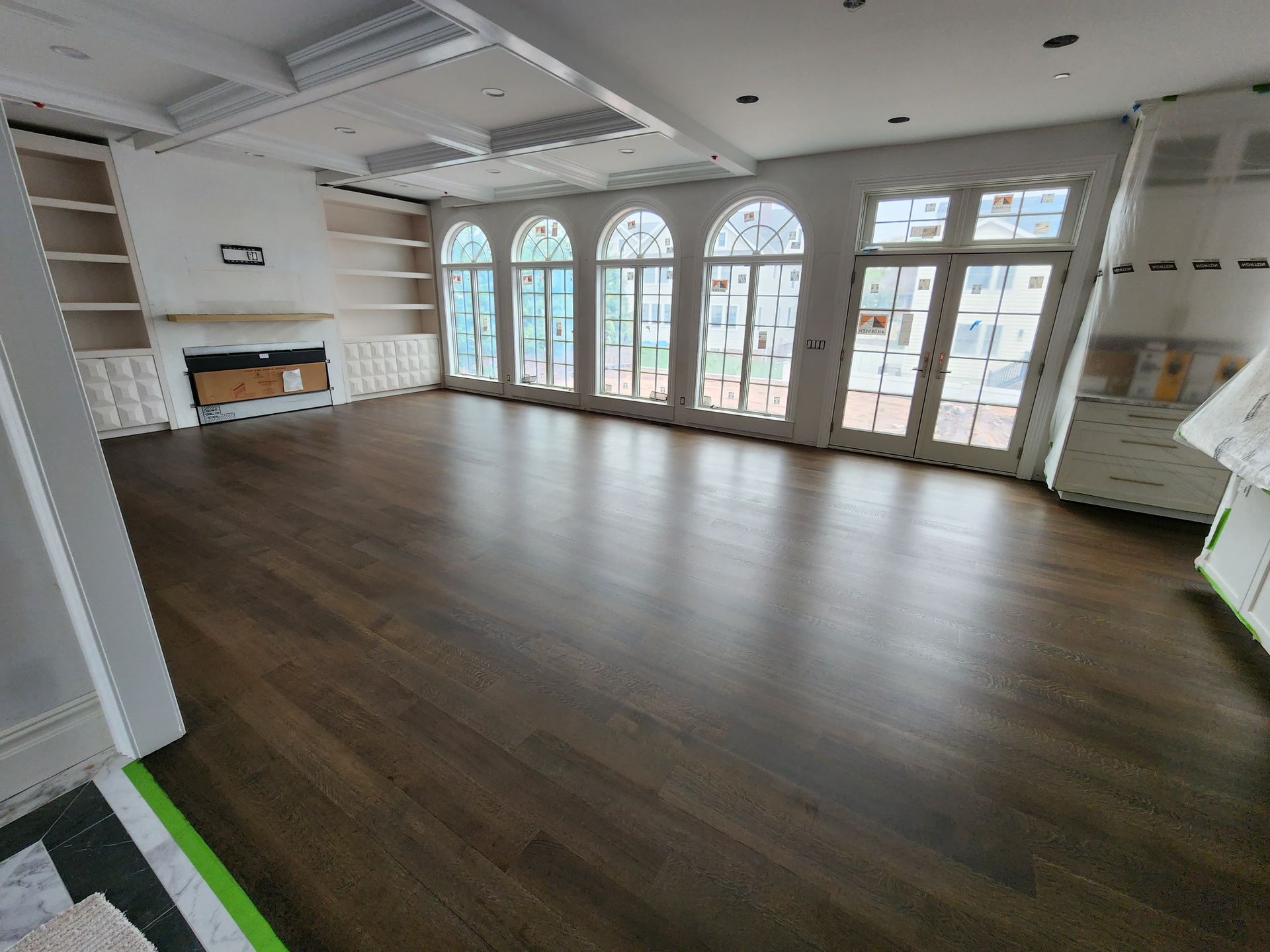 A spacious, bright room with dark wood floors, arched windows, built-in shelving, and a modern fireplace under construction.