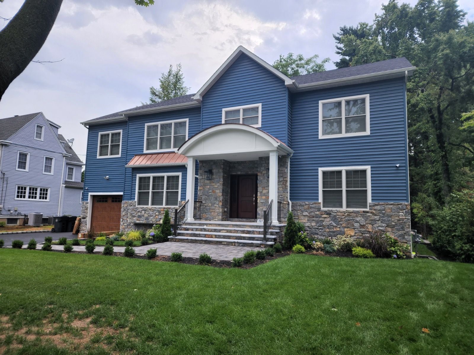 A two-story blue house with stone veneer, a front portico, and a landscaped front lawn on a bright, partly cloudy day.