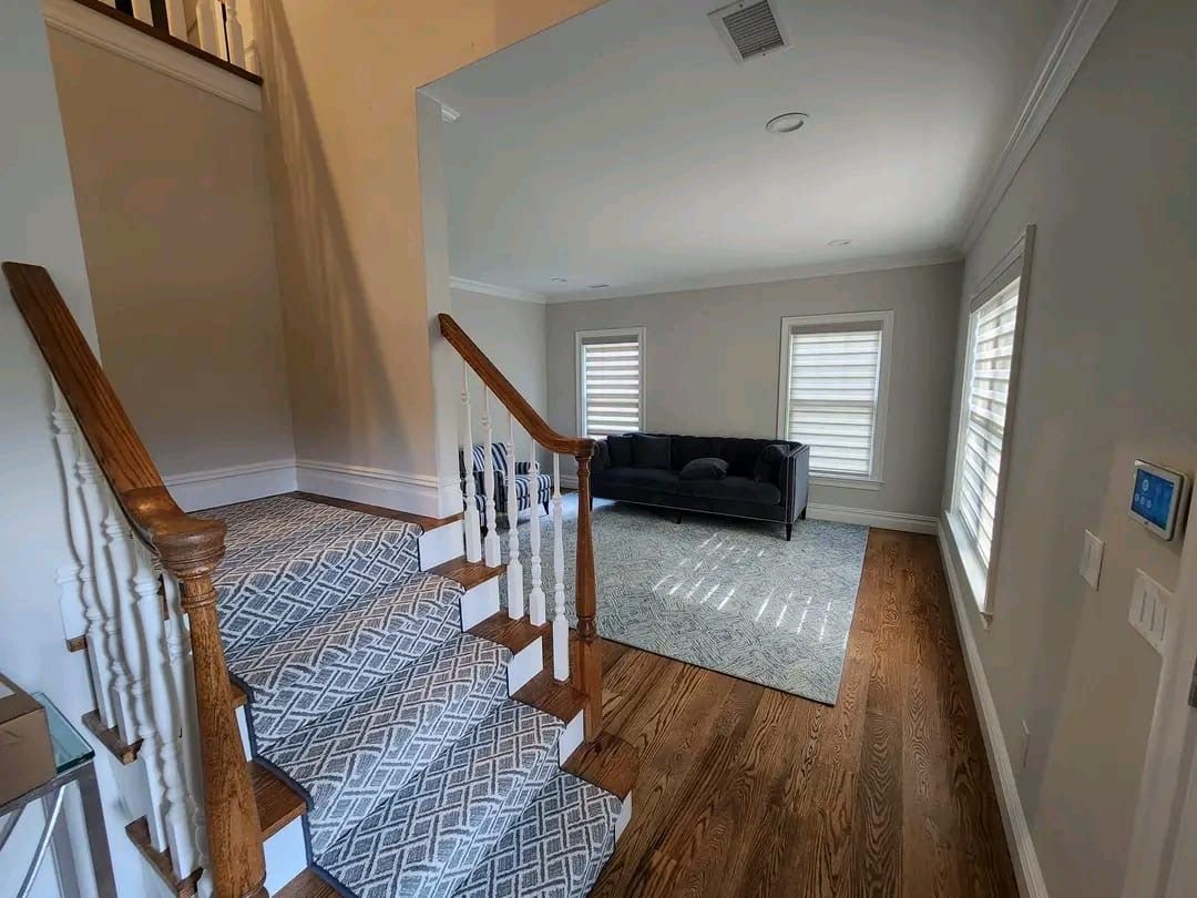 Stairs with patterned carpet lead up from a wood-floored entryway to a bright living room with a dark sofa and area rug.