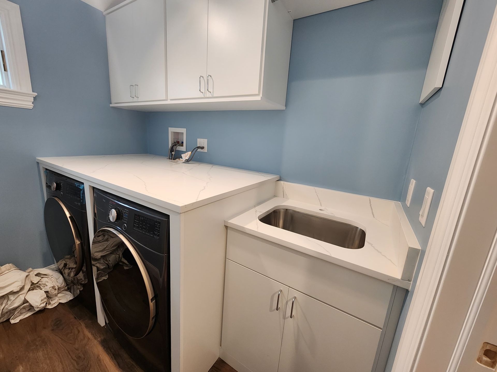 Laundry room with white cabinets, dark washing machines, a sink in a white countertop, and light blue walls.