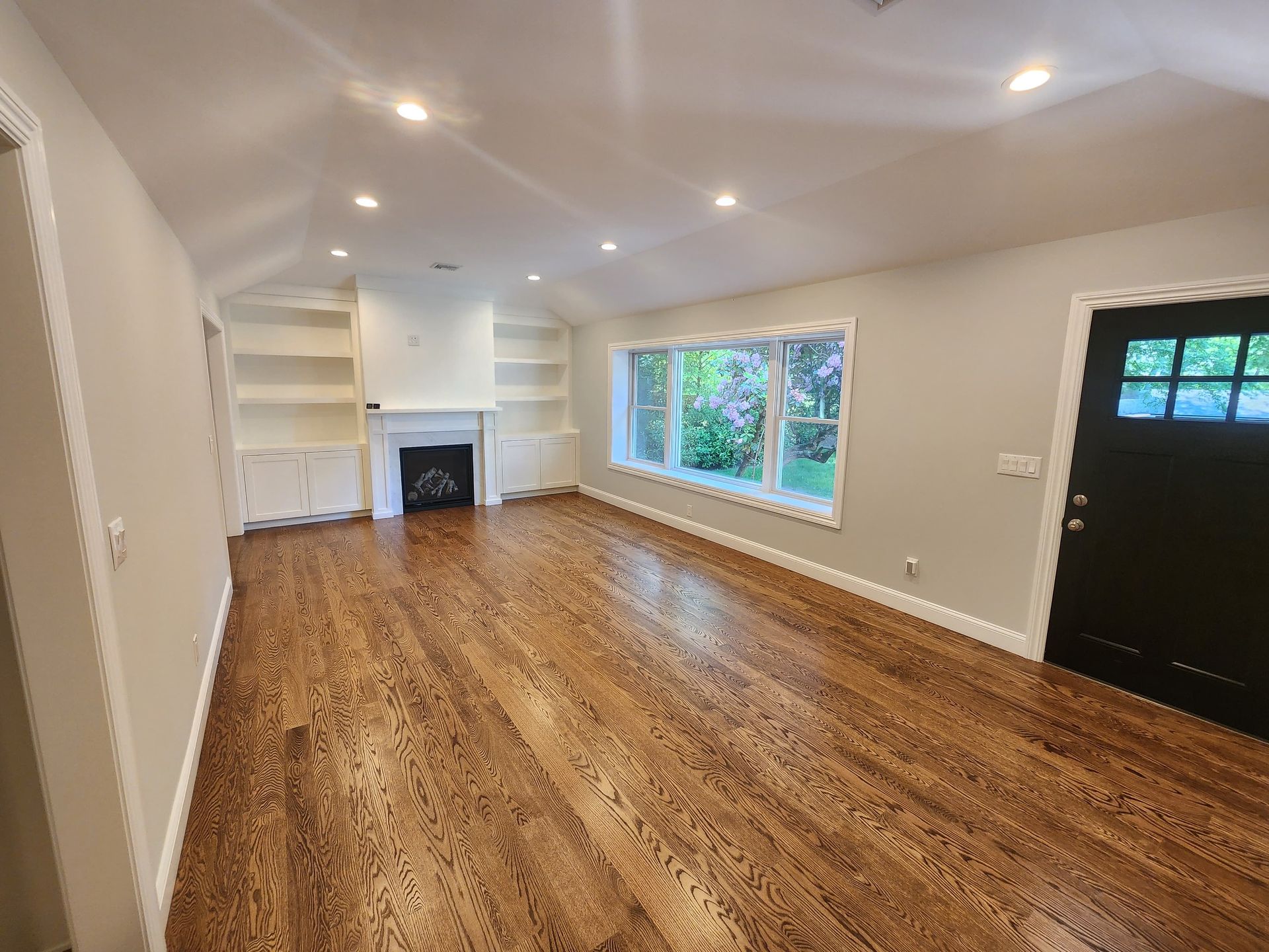 A bright, empty living room with light gray walls, dark wood floors, a fireplace with built-in shelves, and a black door.