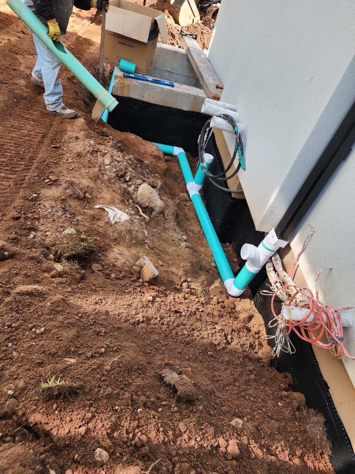 A person installing light green PVC drainage pipes along the foundation of a house under construction.