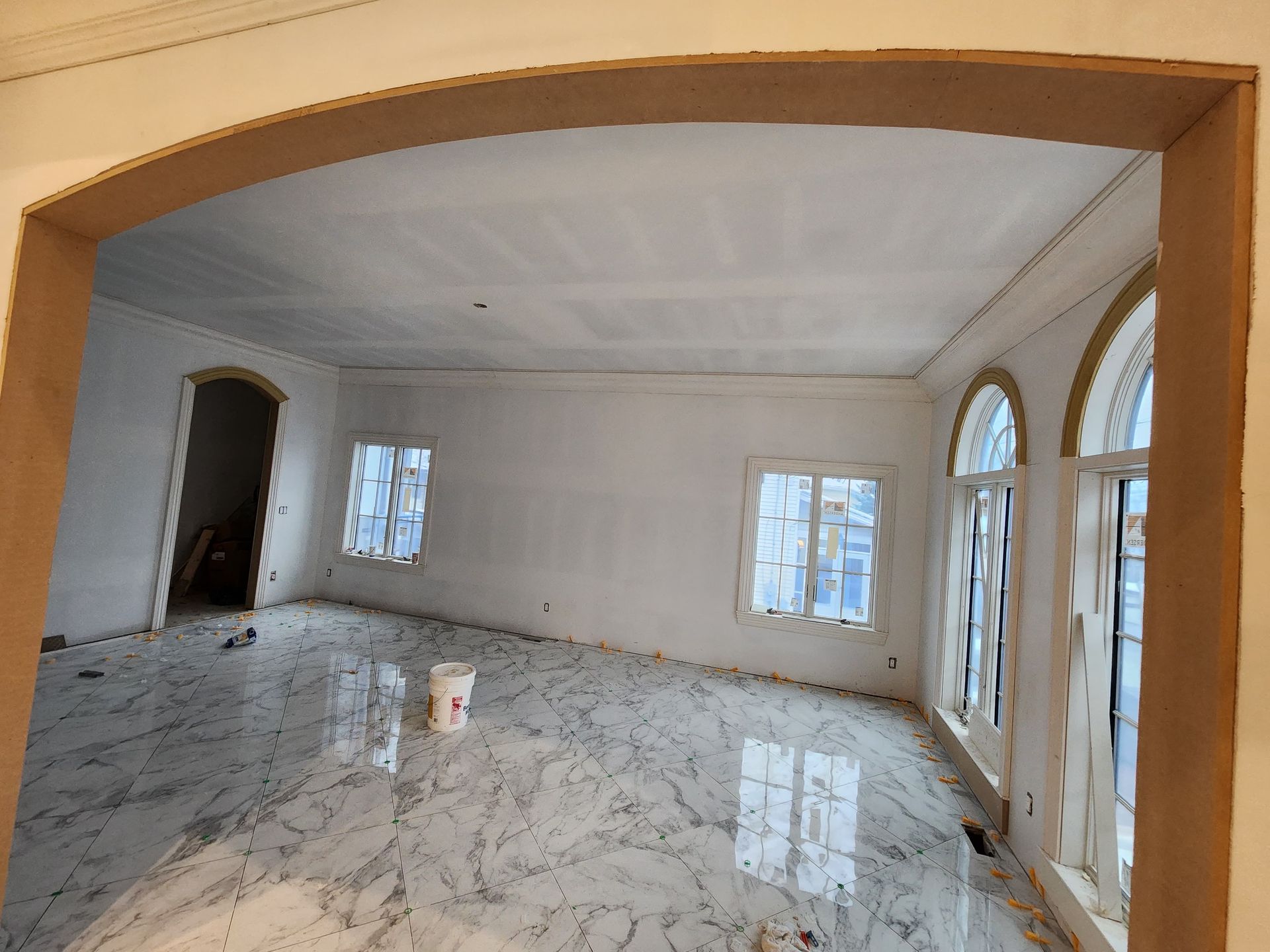 An unfinished room with white walls, arched doorways, marble-patterned tile flooring, and large windows.
