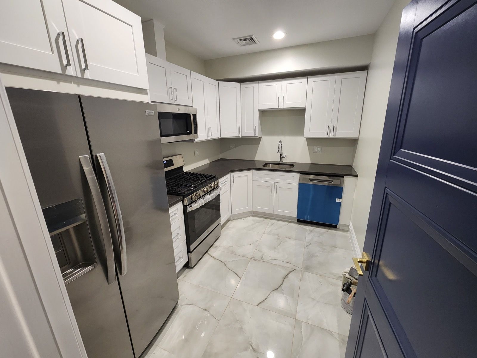 A galley kitchen with white cabinets, stainless steel appliances, dark countertops, and light marble-patterned flooring.