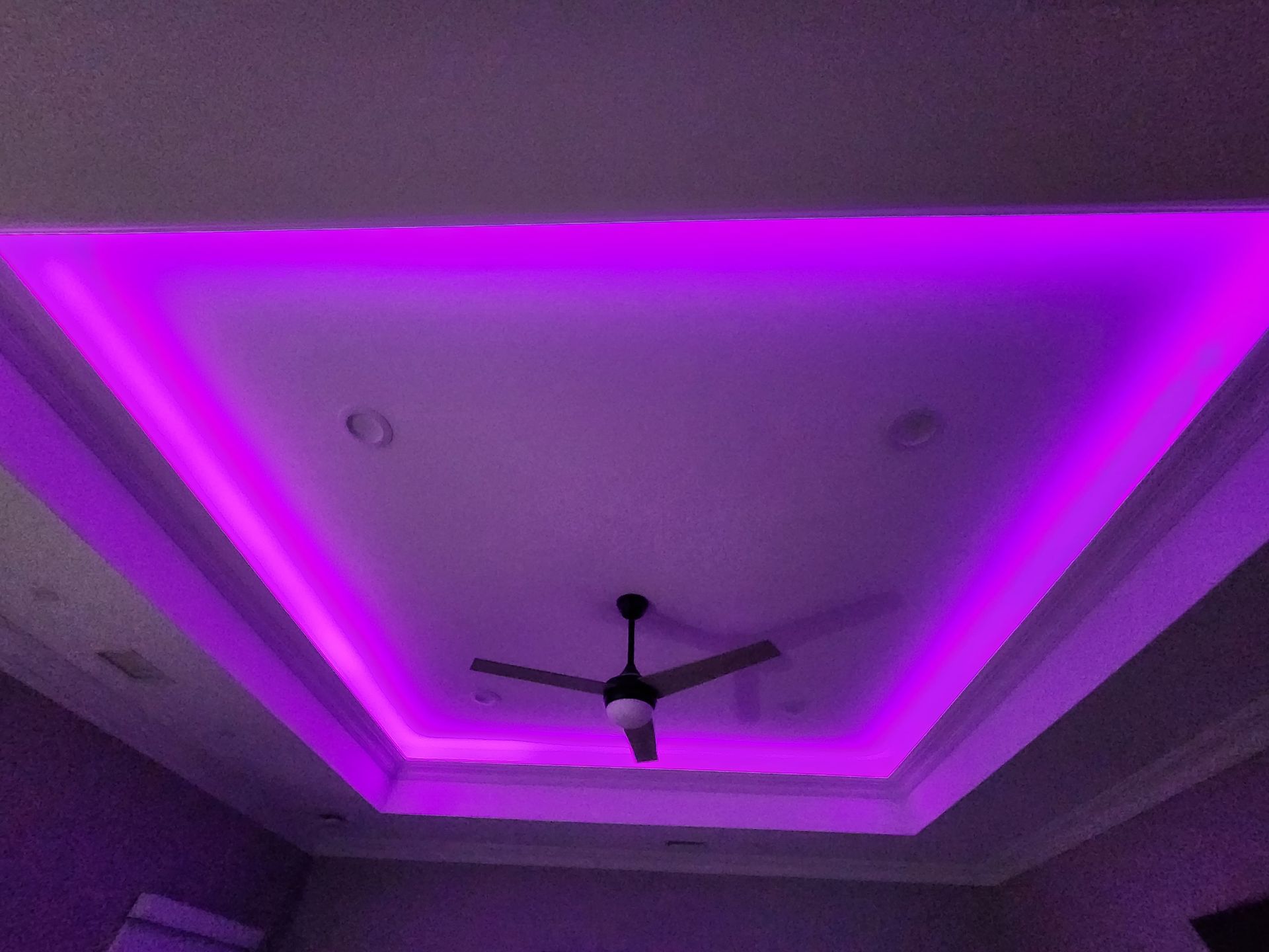 A ceiling with recessed lighting glowing in vibrant purple, featuring a modern ceiling fan in the center.