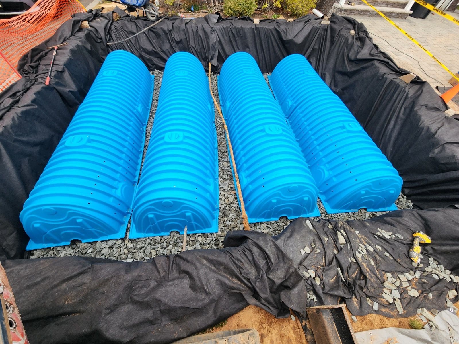 Four bright blue corrugated plastic stormwater drainage chambers installed in a gravel-lined trench with black liners.