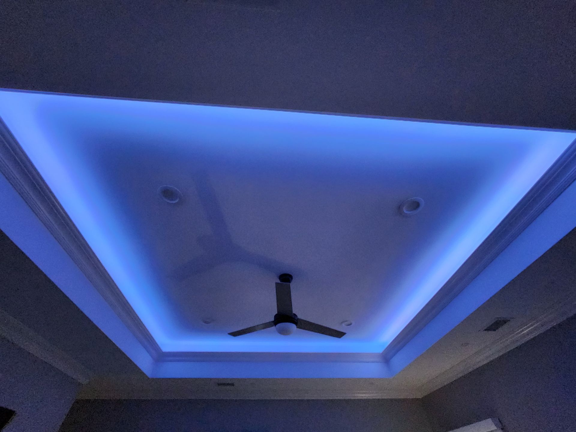 A ceiling with a recessed tray design illuminated by blue LED strip lights and a ceiling fan in the center.
