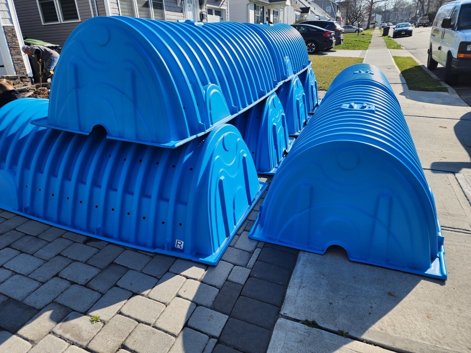 Several bright blue plastic arched storm chamber covers stacked on a paved driveway next to a sidewalk.