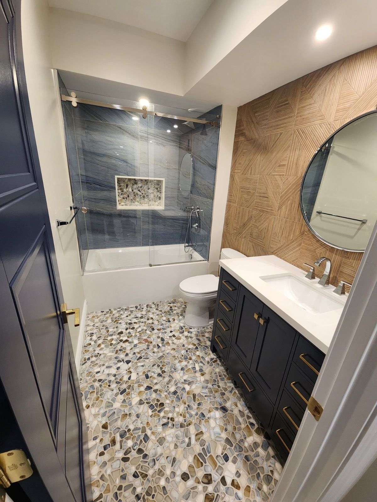 A modern bathroom with a navy blue vanity, gold hardware, a circular mirror, and multi-colored stone-tiled flooring.