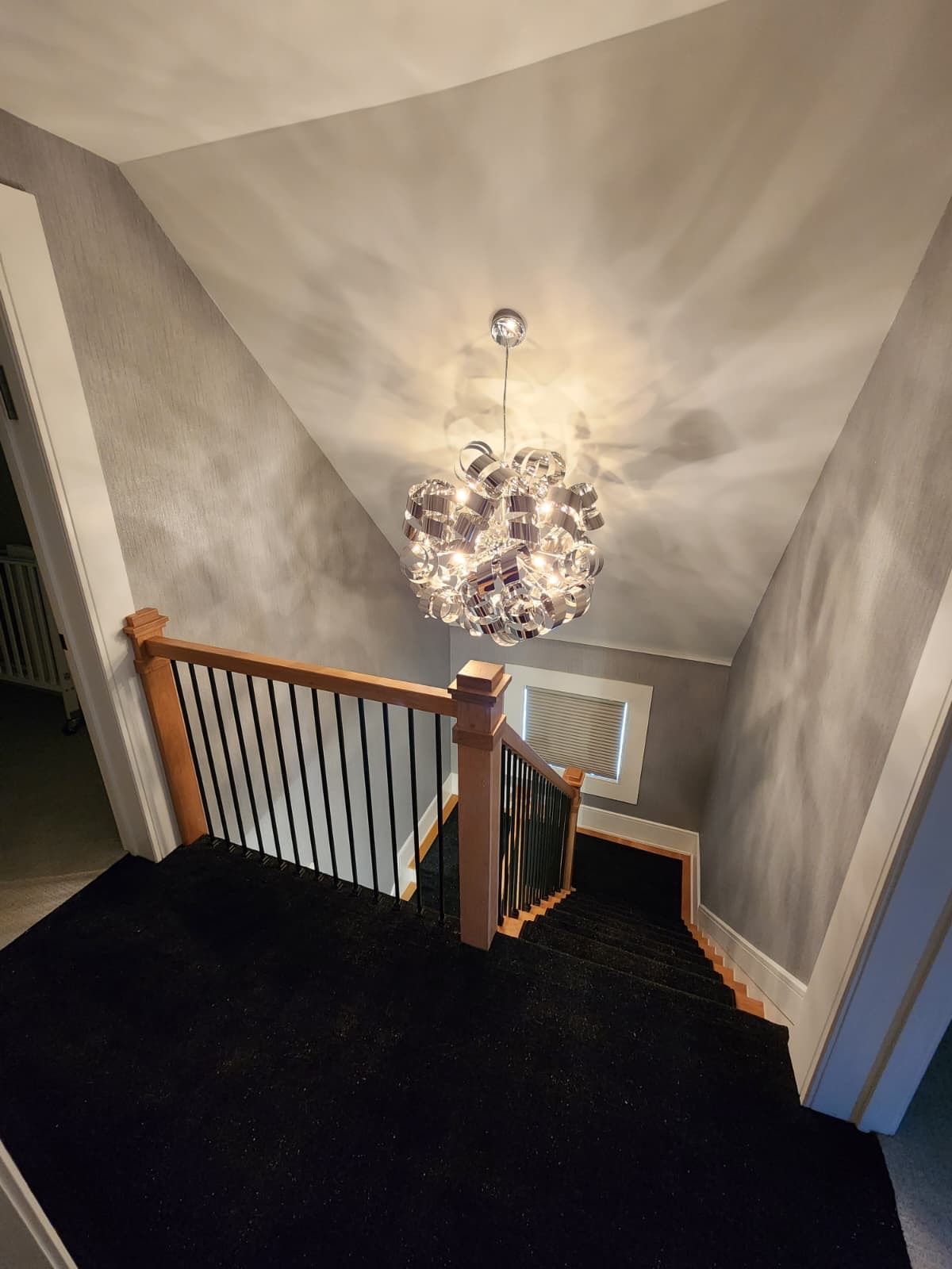A modern, spherical glass chandelier hangs above a staircase with black carpeting, wooden railings, and gray walls.