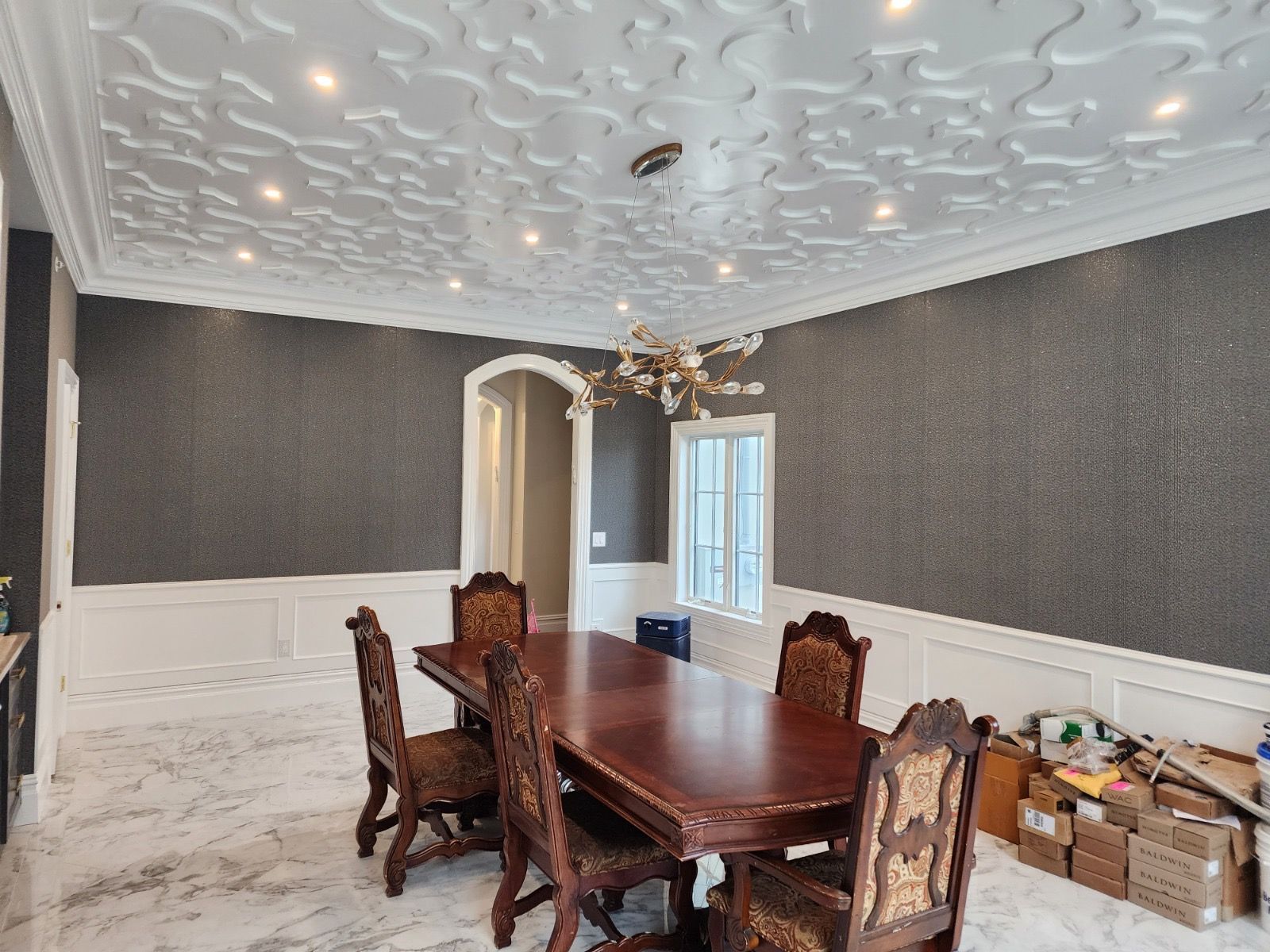 A dining room with a patterned ceiling, dark textured walls, white wainscoting, a wood table, and chairs on marble floors.