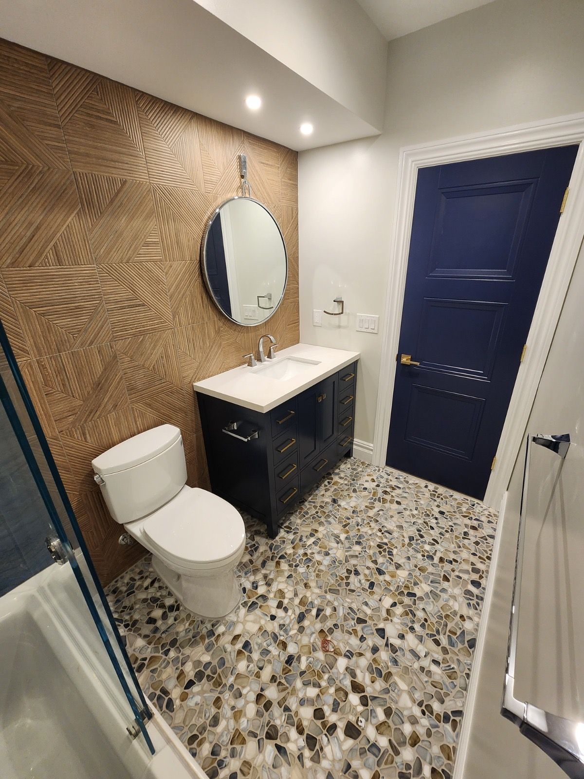 A modern bathroom with textured brown wall, navy vanity, white toilet, a round mirror, and speckled tile floor.