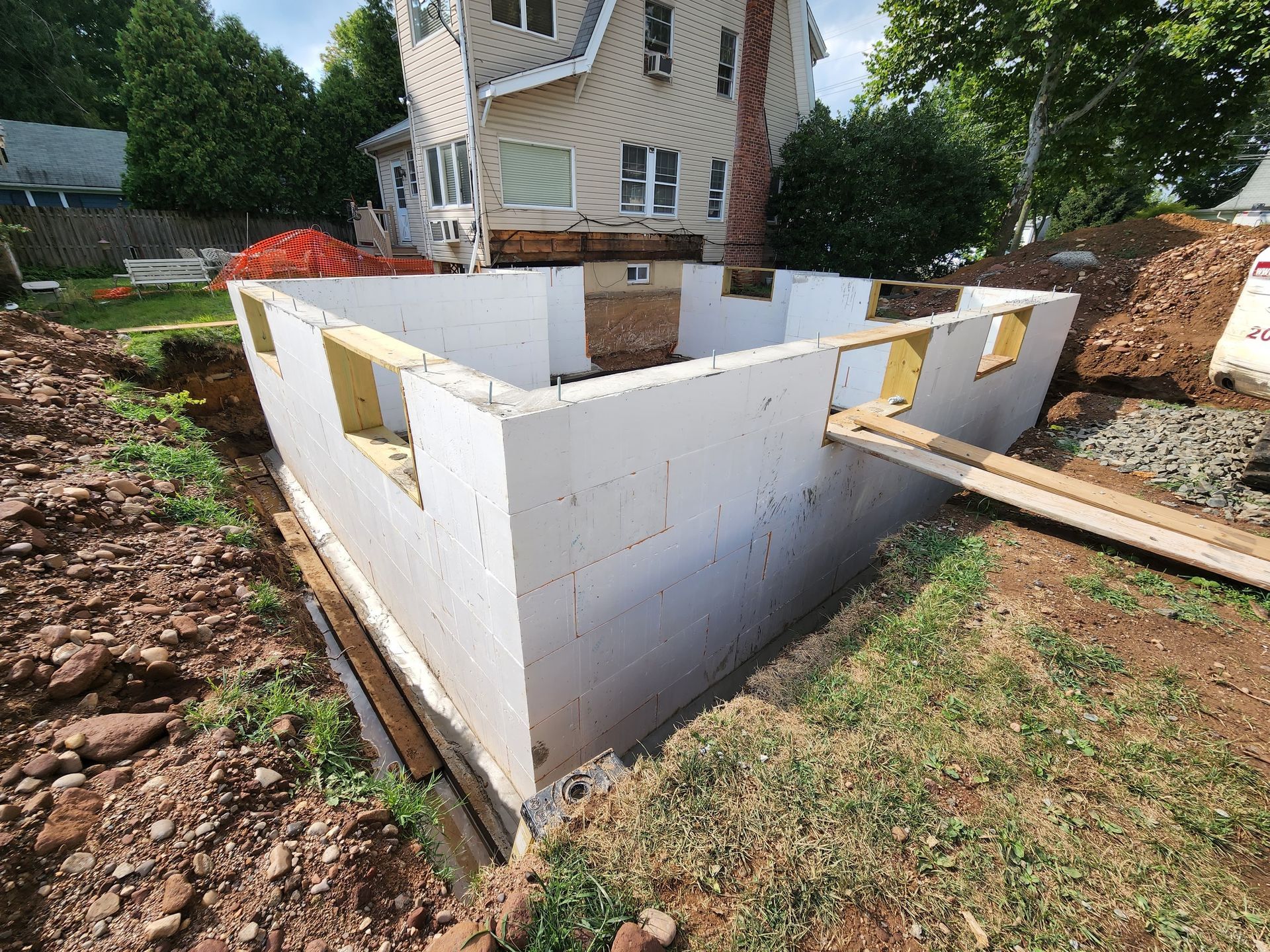 A residential addition foundation made of white insulated concrete forms set in an excavated yard beside a house.
