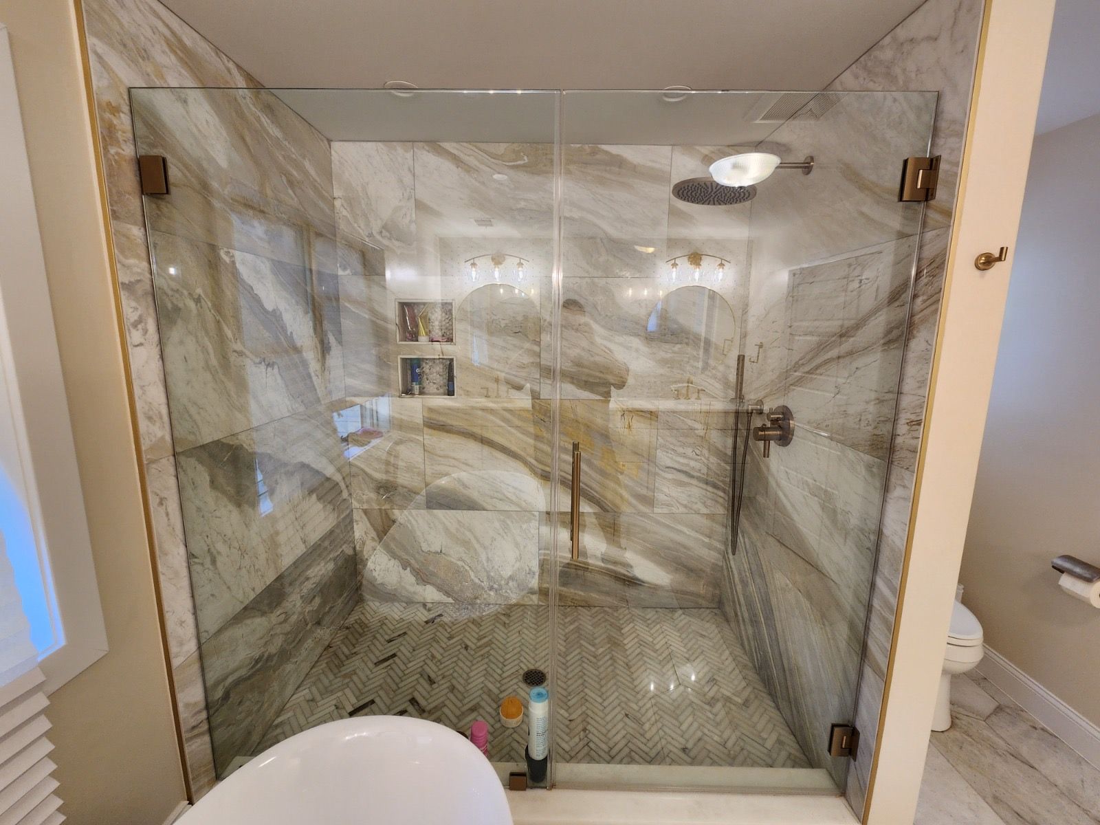 A walk-in shower with gold-trimmed glass doors, light-veined marble walls, a rainfall showerhead, and a tiled floor.