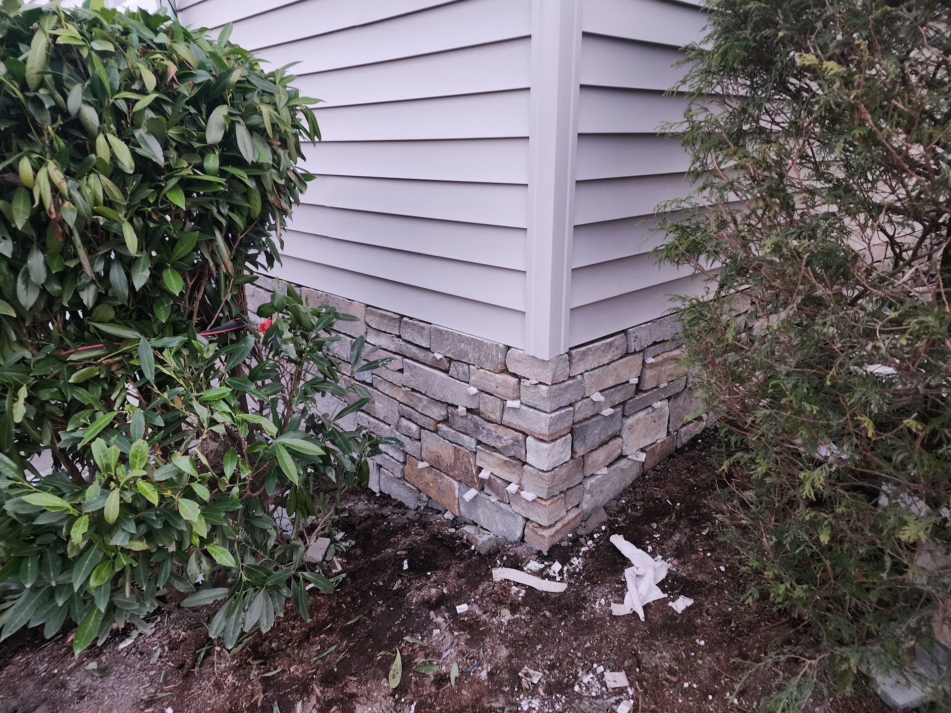 The exterior corner of a house featuring gray vinyl siding above a stone veneer foundation, flanked by green bushes.