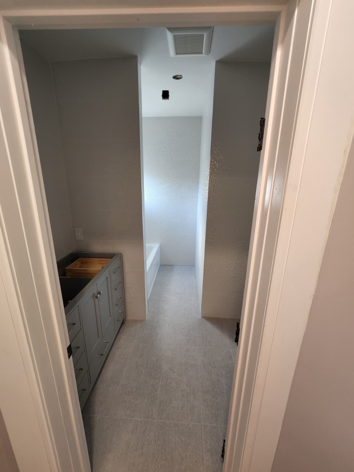 A narrow bathroom featuring a gray vanity on the left, light-colored tile flooring, and a bathtub at the far end.