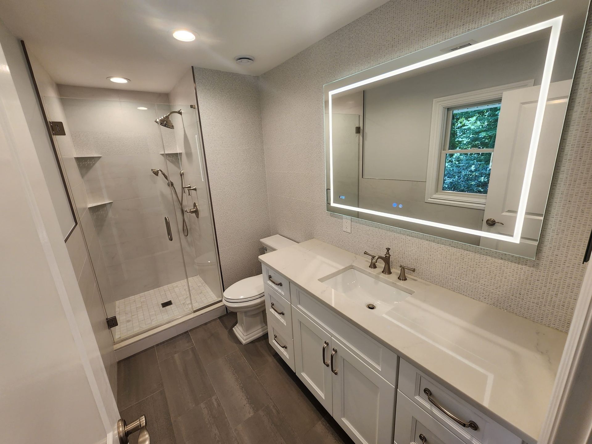 Modern bathroom with a glass-enclosed shower, a white vanity, a large illuminated mirror, and wood-look flooring.