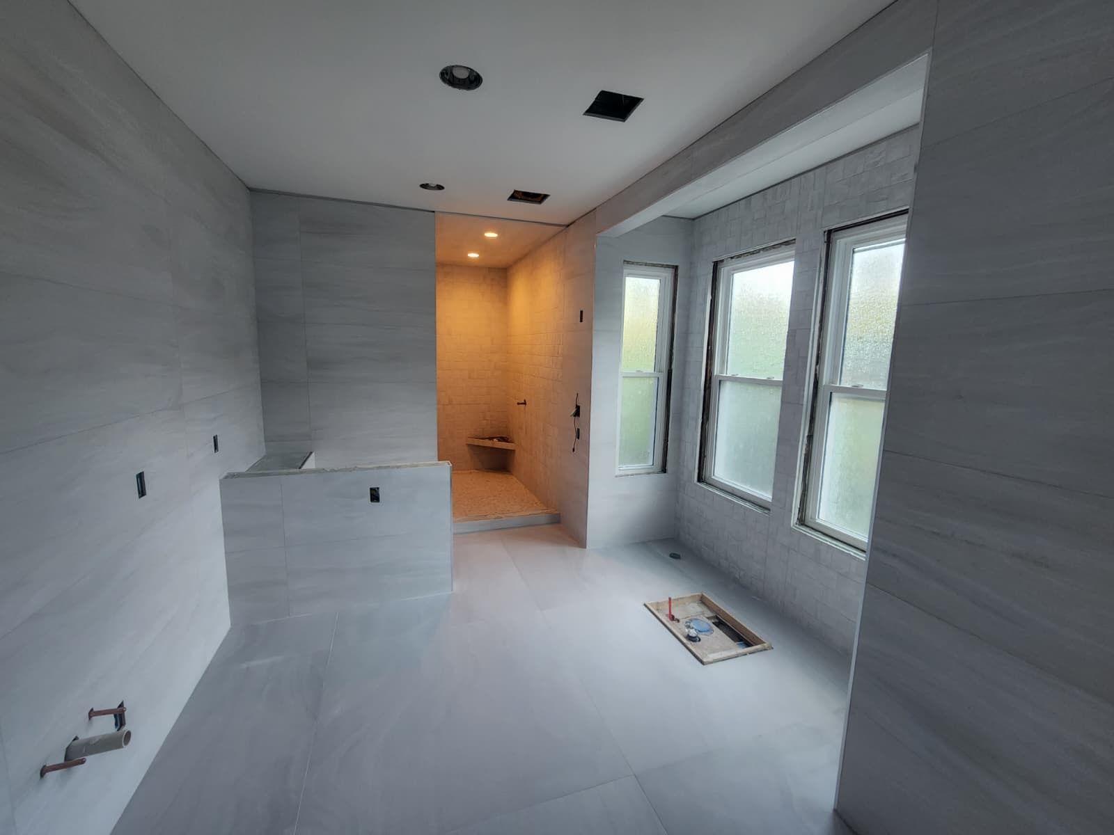 A wide shot of a modern bathroom under construction, featuring large light gray wall tiles, a soaking tub, and a shower area.