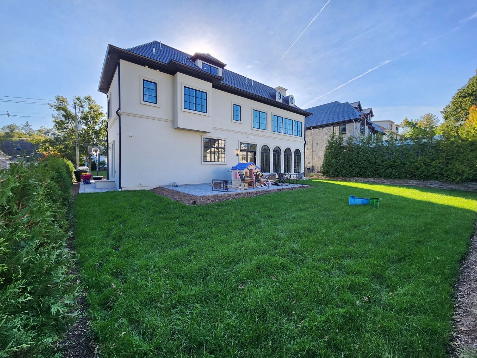 A two-story light-colored house with a dark roof and backyard lawn, bordered by tall green hedges on a sunny day.