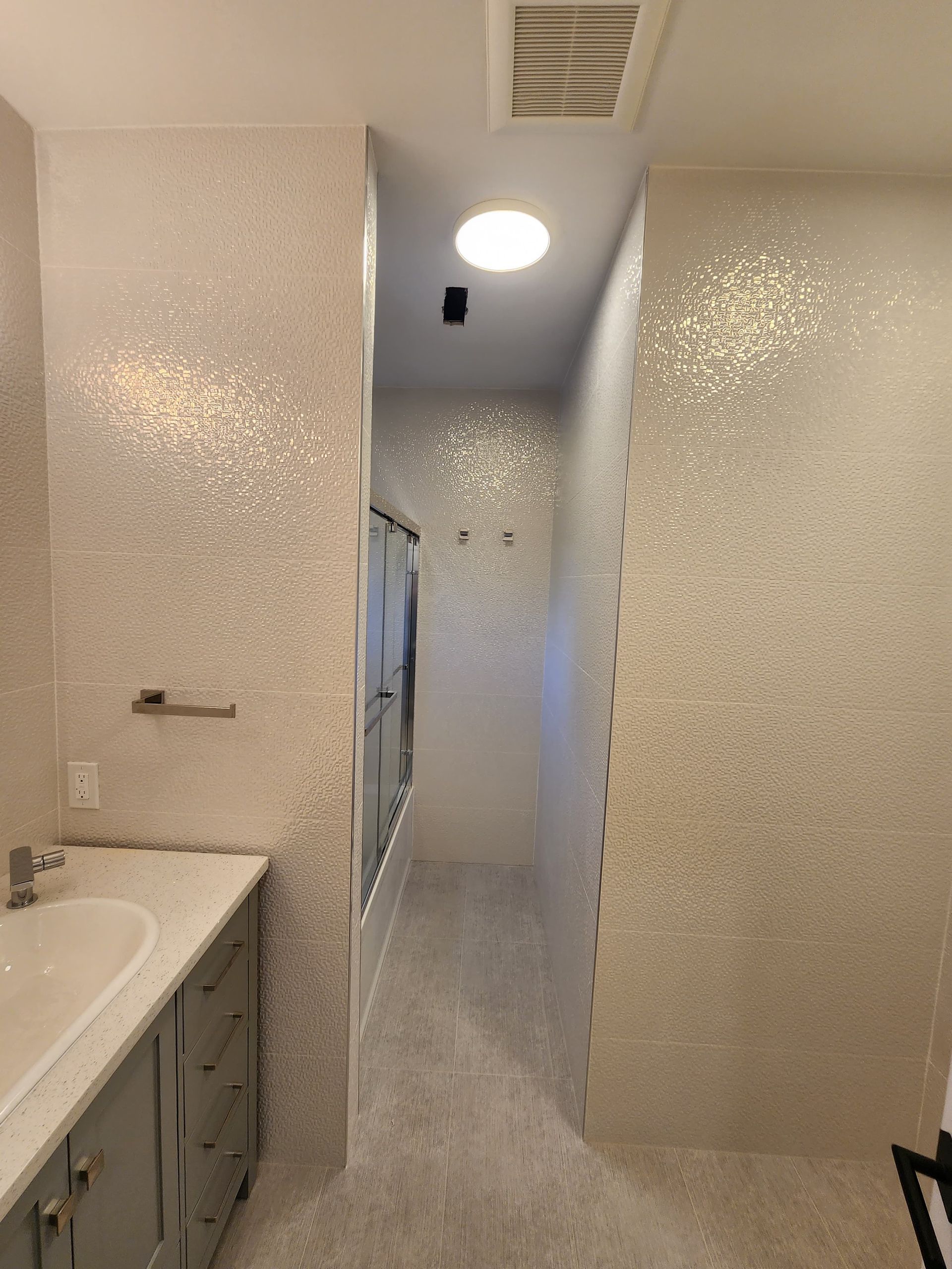 A bathroom with grey vanity, white marble countertop, and walls covered in light-colored, reflective, mosaic-style tiles.