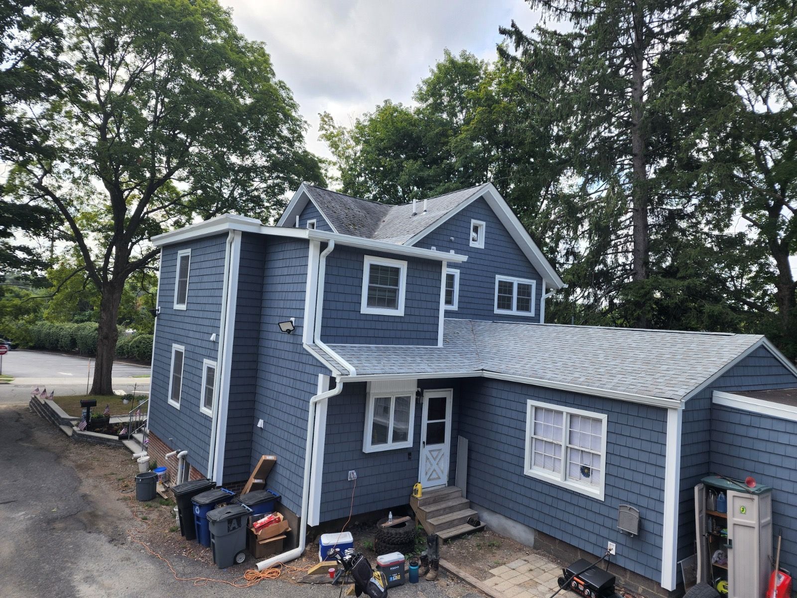 A two-story blue house with white trim and a shingled roof, viewed from an elevated angle on a cloudy day.