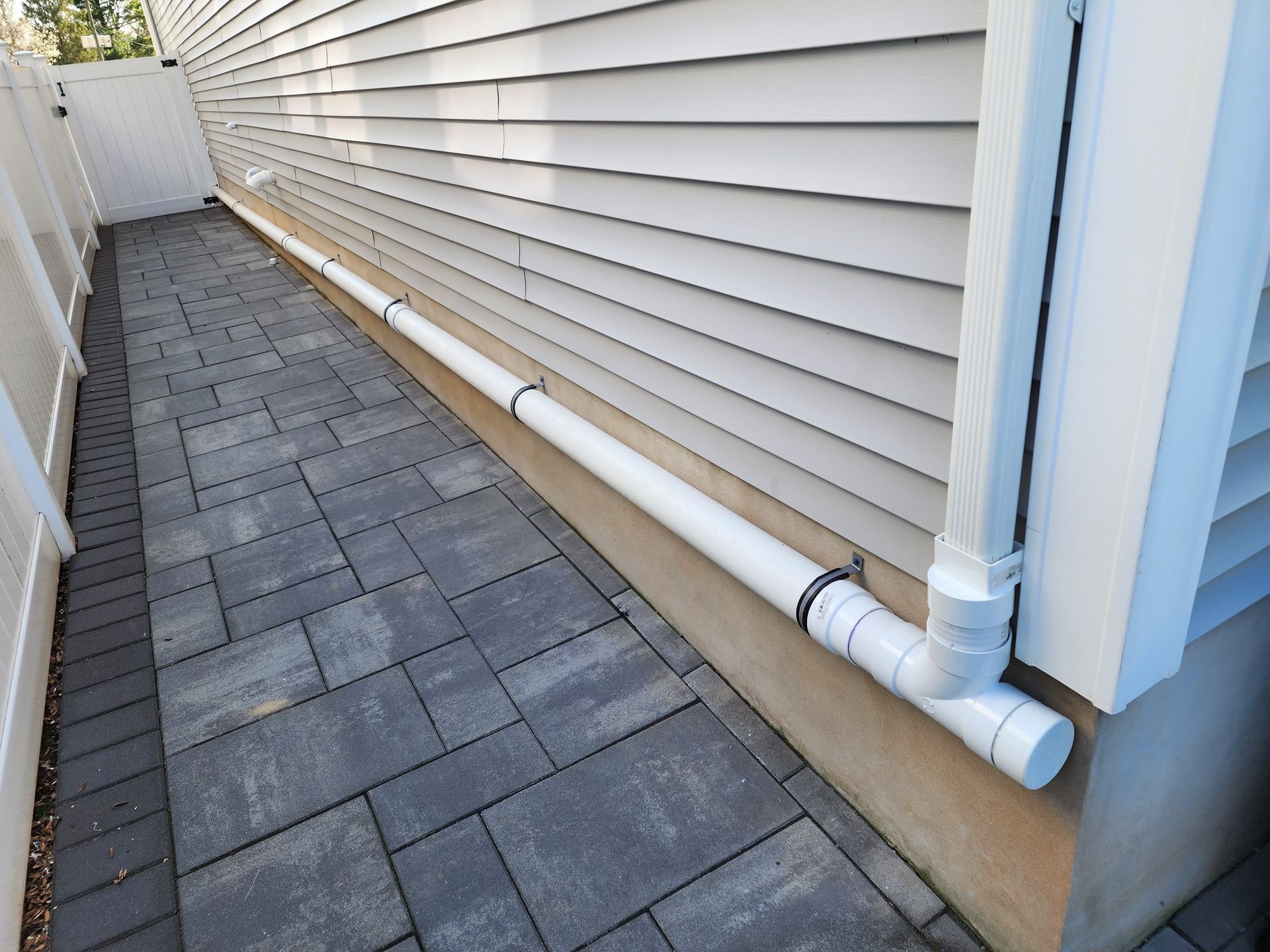 A white PVC downspout extension runs along a house wall, directing water onto a gray paved walkway.