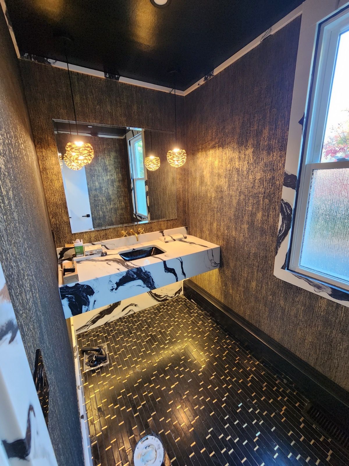 Modern bathroom with marble-patterned vanity, black-and-gold textured wallpaper, hanging globe lights, and tiled floor.
