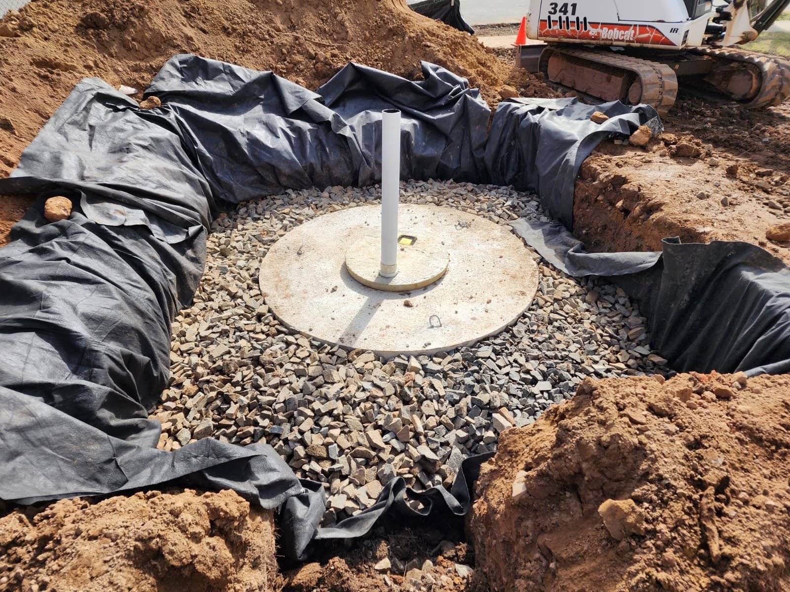 A circular concrete structure with a vertical white pipe sits in a gravel-filled, black-lined trench during construction.